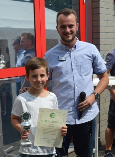 Junge h&auml;lt eine Ehrenurkunde und einen Pokal, w&auml;hrend ein Mann mit Mikrofon neben ihm steht. Hintergrund mit weiteren Personen.