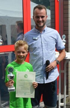 Junger Sportler in gr&uuml;ner T-Shirt h&auml;lt Pokal und Urkunde, w&auml;hrend er neben einem Erwachsenen mit Mikrofon steht.