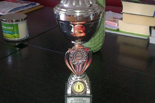 Silberner Pokal mit &bdquo;2017&ldquo;-Plakette und zwei Geschenkgutscheinen auf einem Tisch, im Hintergrund eine Blumenvase.