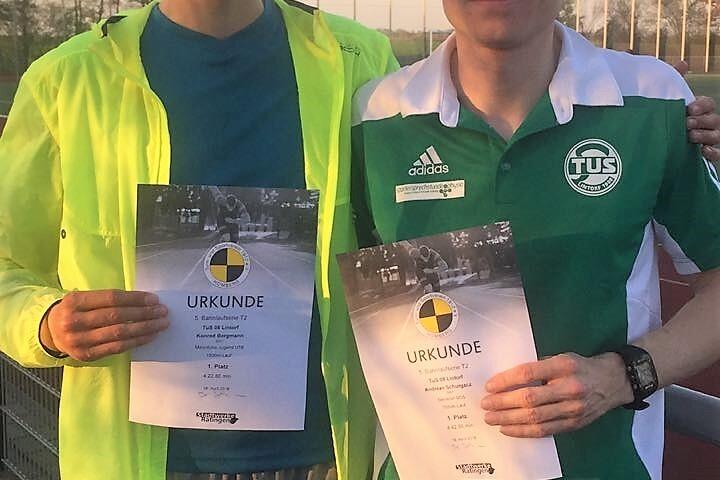 Zwei M&auml;nner halten Urkunden in der Hand, stehen l&auml;chelnd auf einem Sportplatz bei Sonnenuntergang.