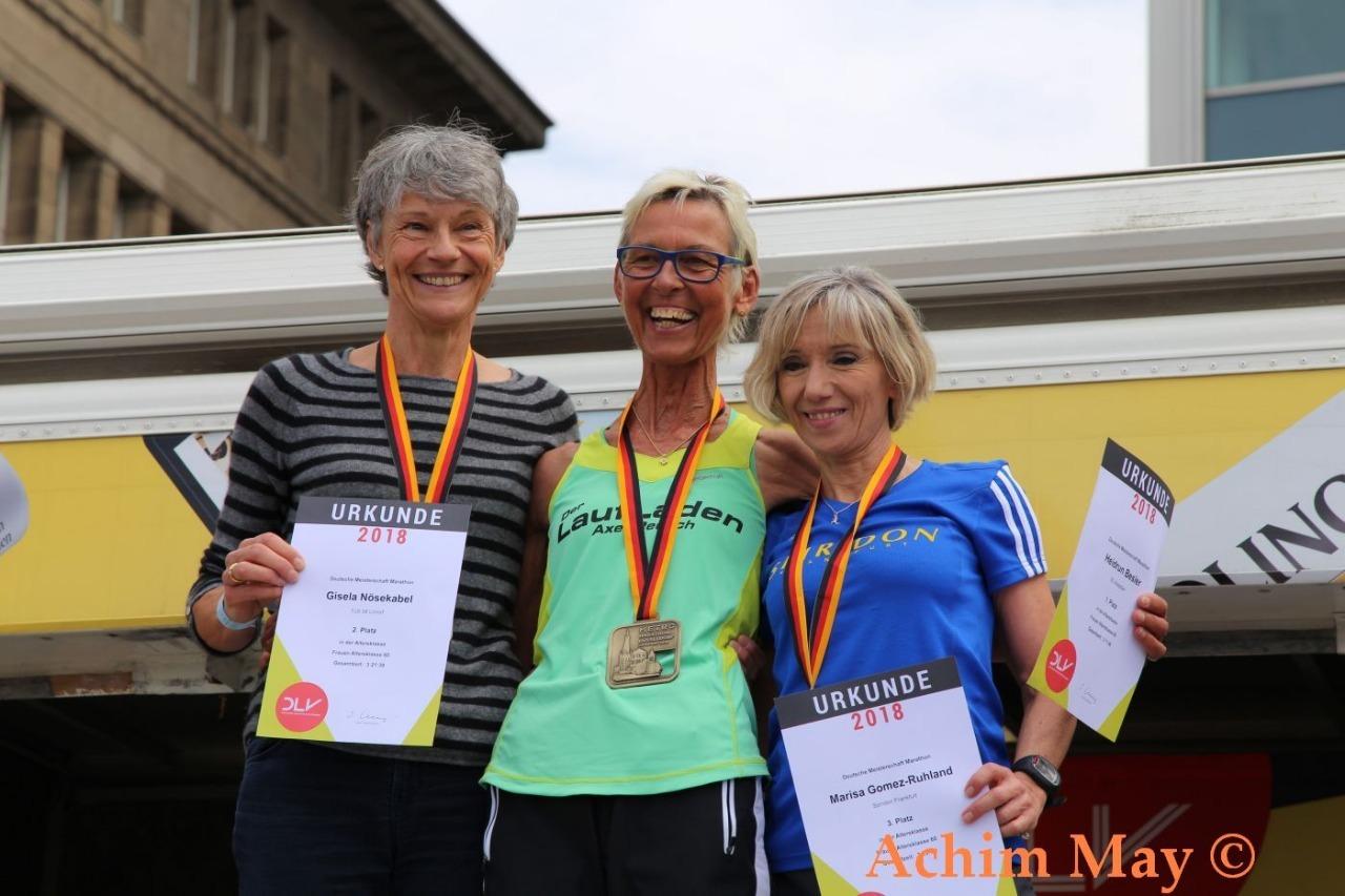 Drei Frauen halten Urkunden und Medaillen bei einer Siegerehrung, l&auml;chelnd auf einer B&uuml;hne mit bunten Medaillen.