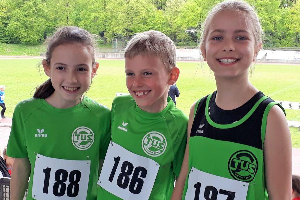 Drei Kinder in grünen Sporttrikots mit Startnummern lächeln in die Kamera vor einem Sportplatz.