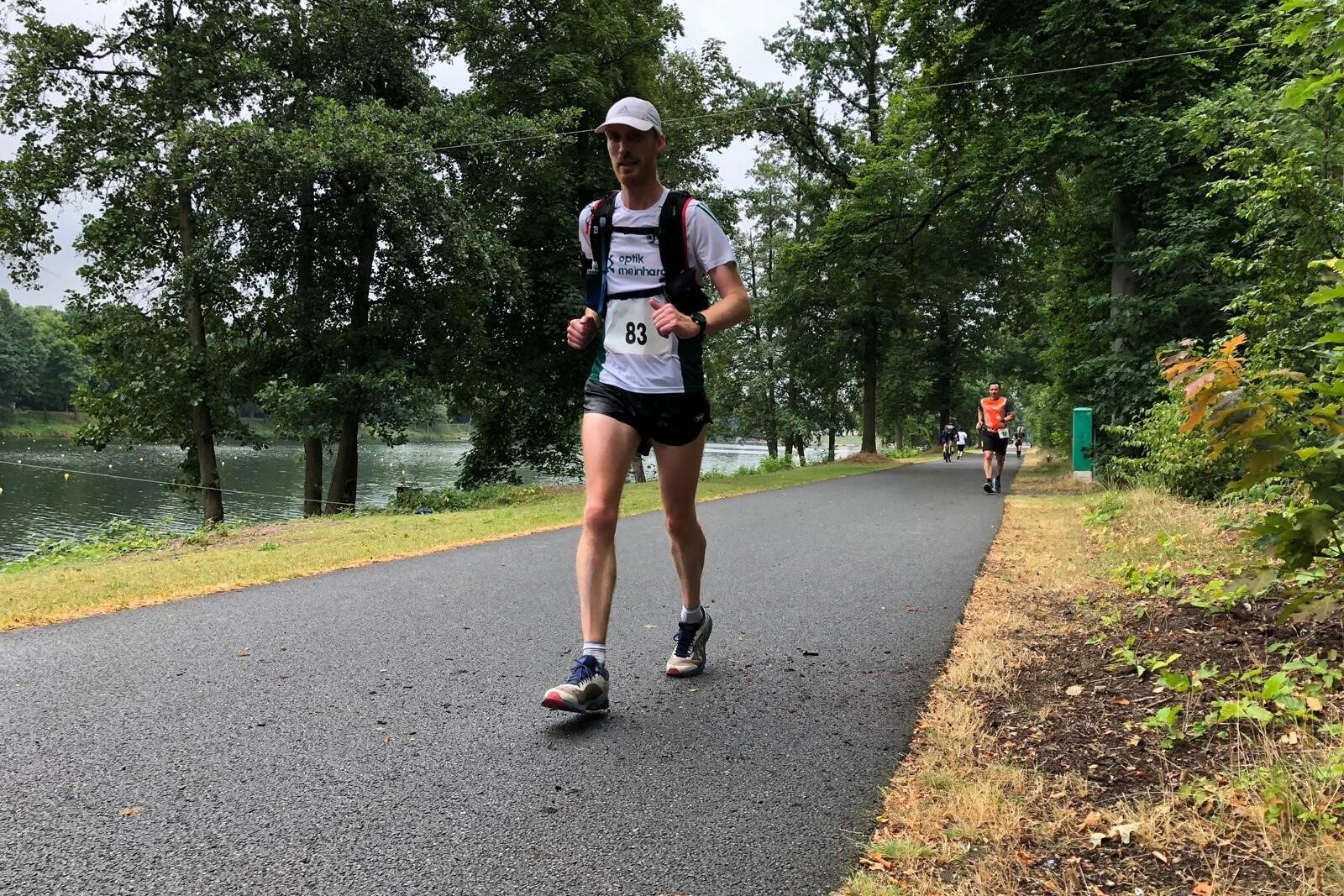 Läufer in sportlicher Kleidung mit Nummer 83 joggt auf einem Weg am See, umgeben von Bäumen und Natur.