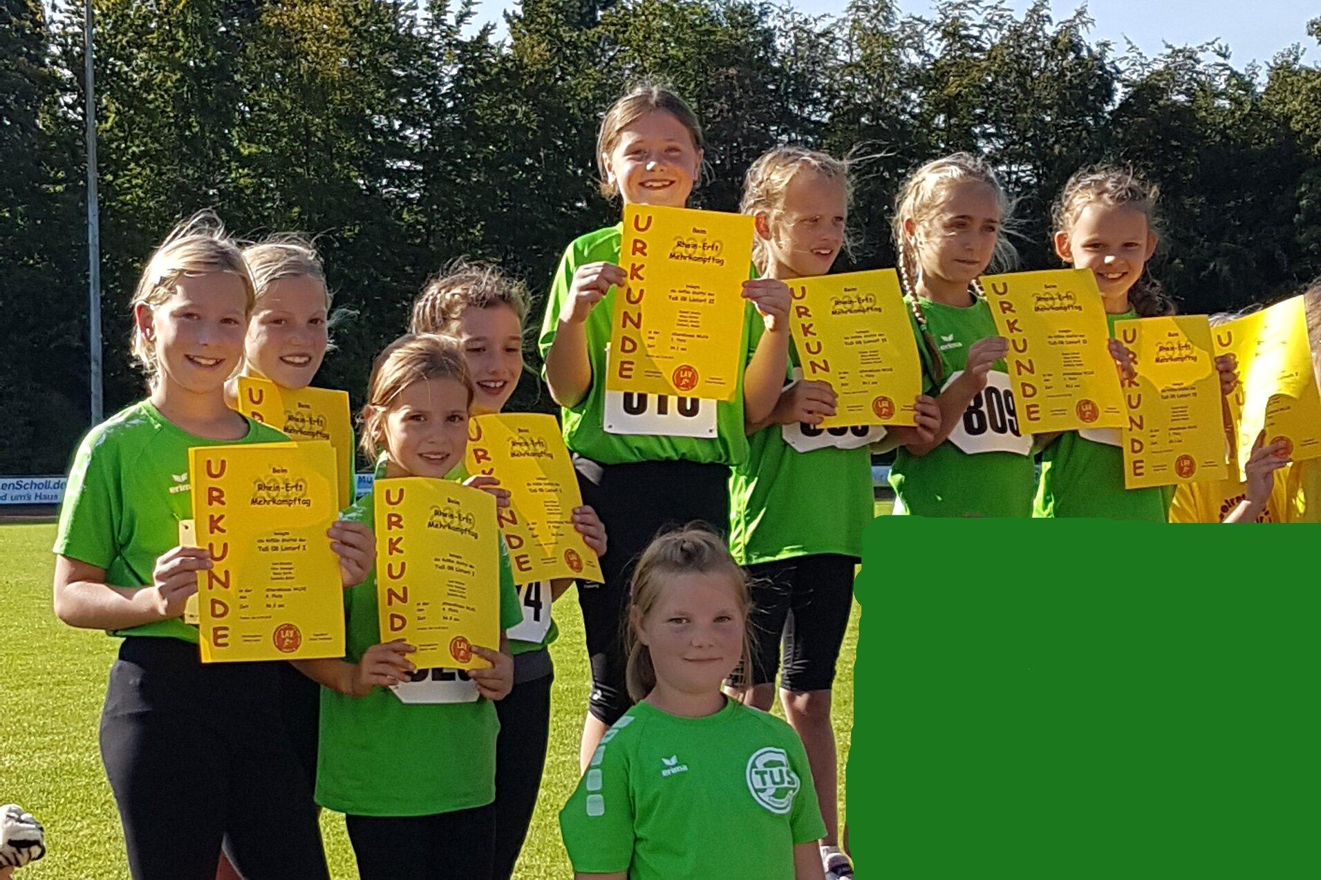 Gruppe von zehn M&auml;dchen in gr&uuml;nen T-Shirts, die stolz ihre Urkunden auf dem Siegerpodest zeigen.