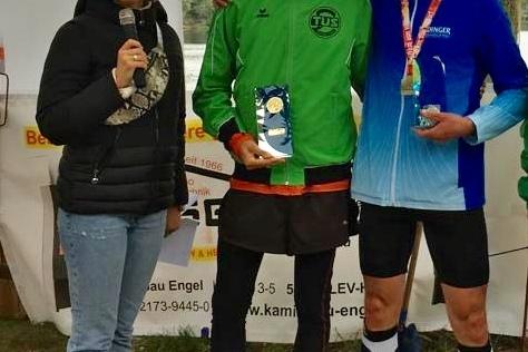 Drei Personen stehen vor einem Banner: Zwei L&auml;ufer in Sportkleidung mit Medaillen, eine Frau h&auml;lt ein Mikrofon.