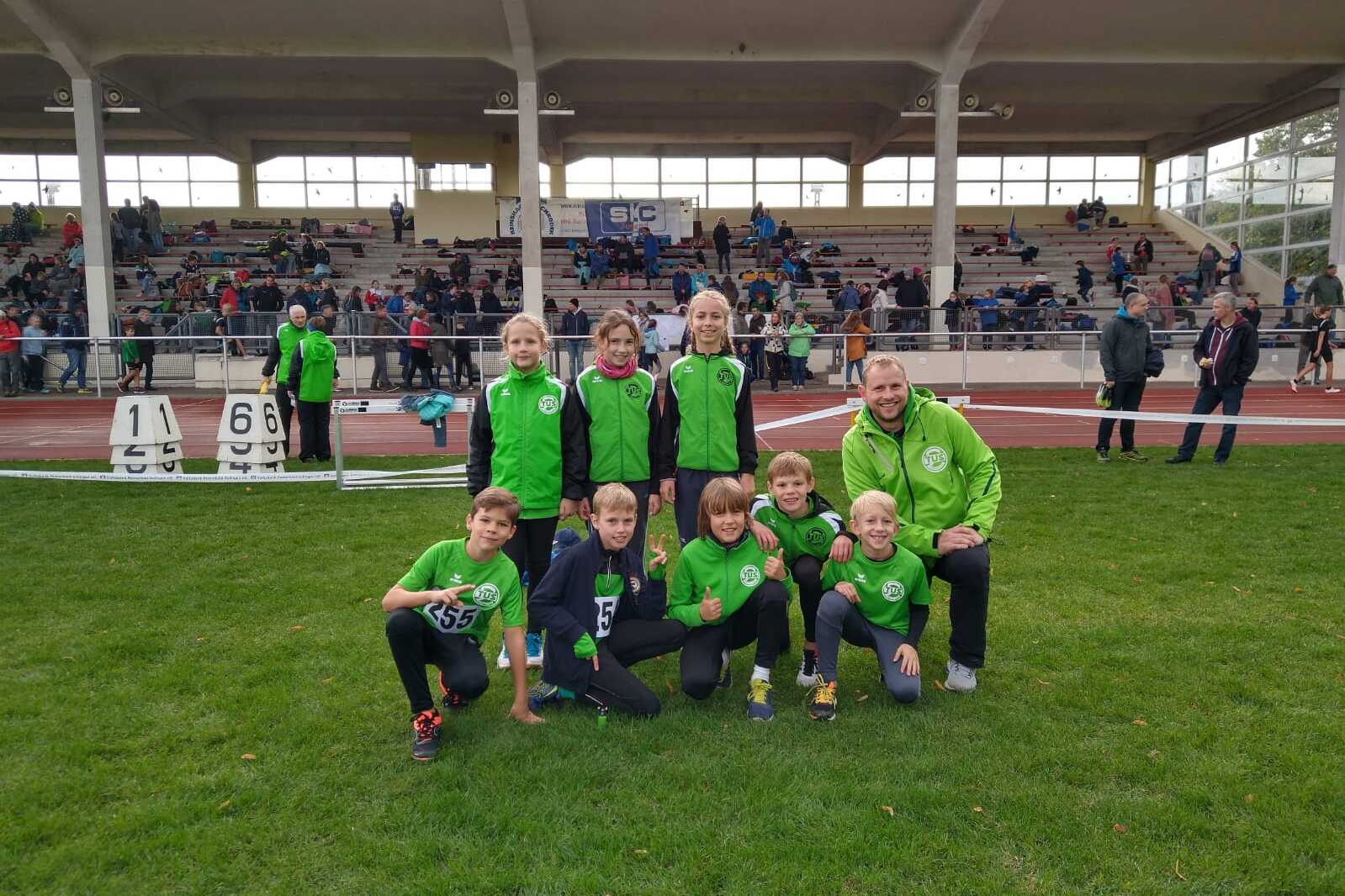 Gruppenfoto von zehn Kindern in gr&uuml;nen Sportanz&uuml;gen, posierend auf einer Laufbahn vor Zuschauertrib&uuml;ne.