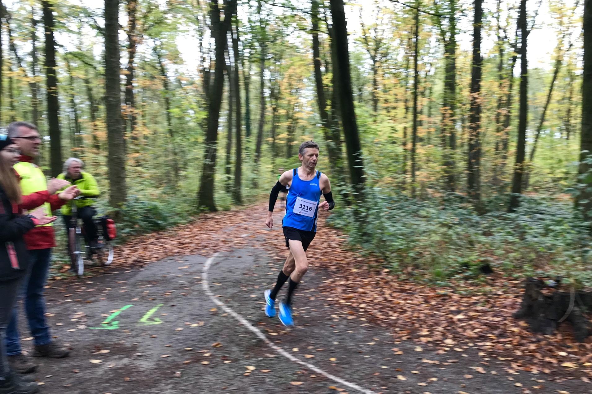 L&auml;ufer in blauer Sportbekleidung sprintet auf einem Waldweg, w&auml;hrend Zuschauer applaudieren. Herbstliche Baumlandschaft im Hintergrund.