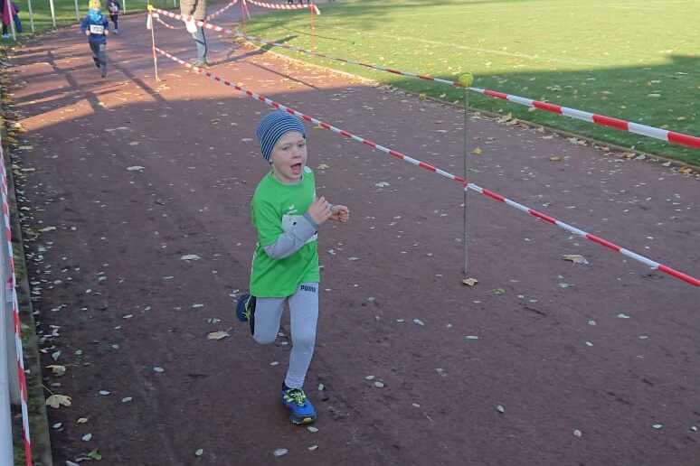 Ein Junge in gr&uuml;ner Sportkleidung l&auml;uft auf einer Laufbahn, umgeben von herbstlichen B&auml;umen und Zuschauern.