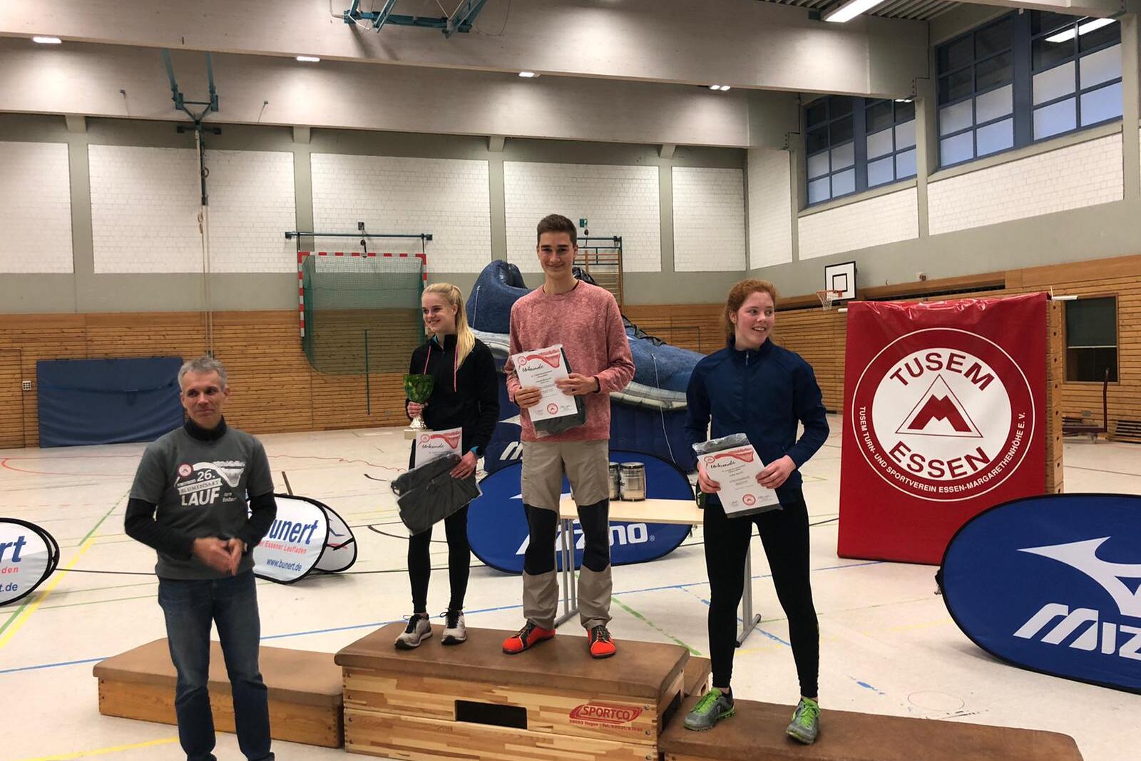 Drei Sportler stehen auf dem Siegertreppchen und halten Urkunden in einer Sporthalle, mit Sponsorenbannern im Hintergrund.