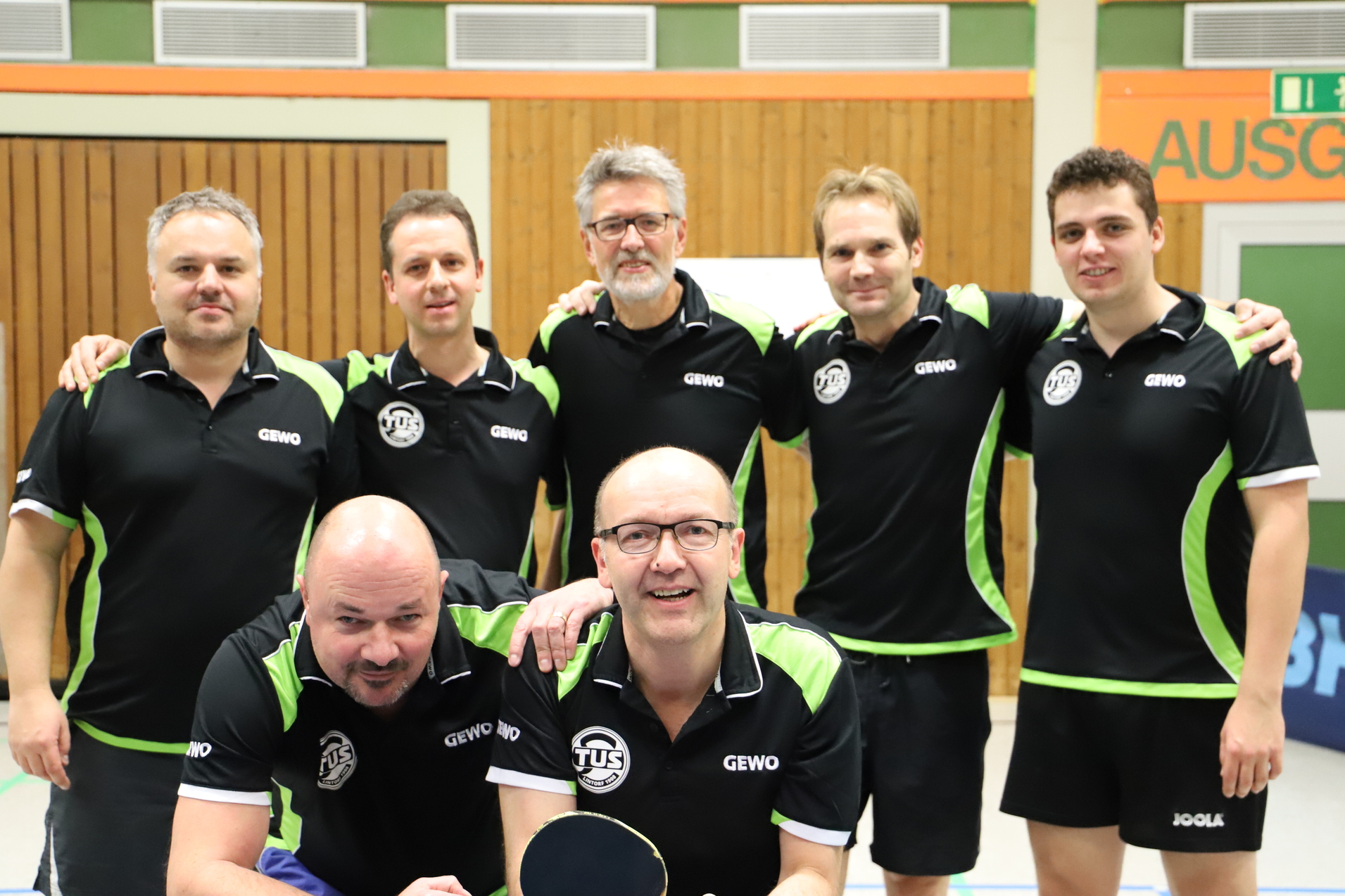 Sechs M&auml;nner in schwarz-gr&uuml;nen Tischtennistrikots posieren freundlich vor einer Tischtennisplatte in einer Halle.
