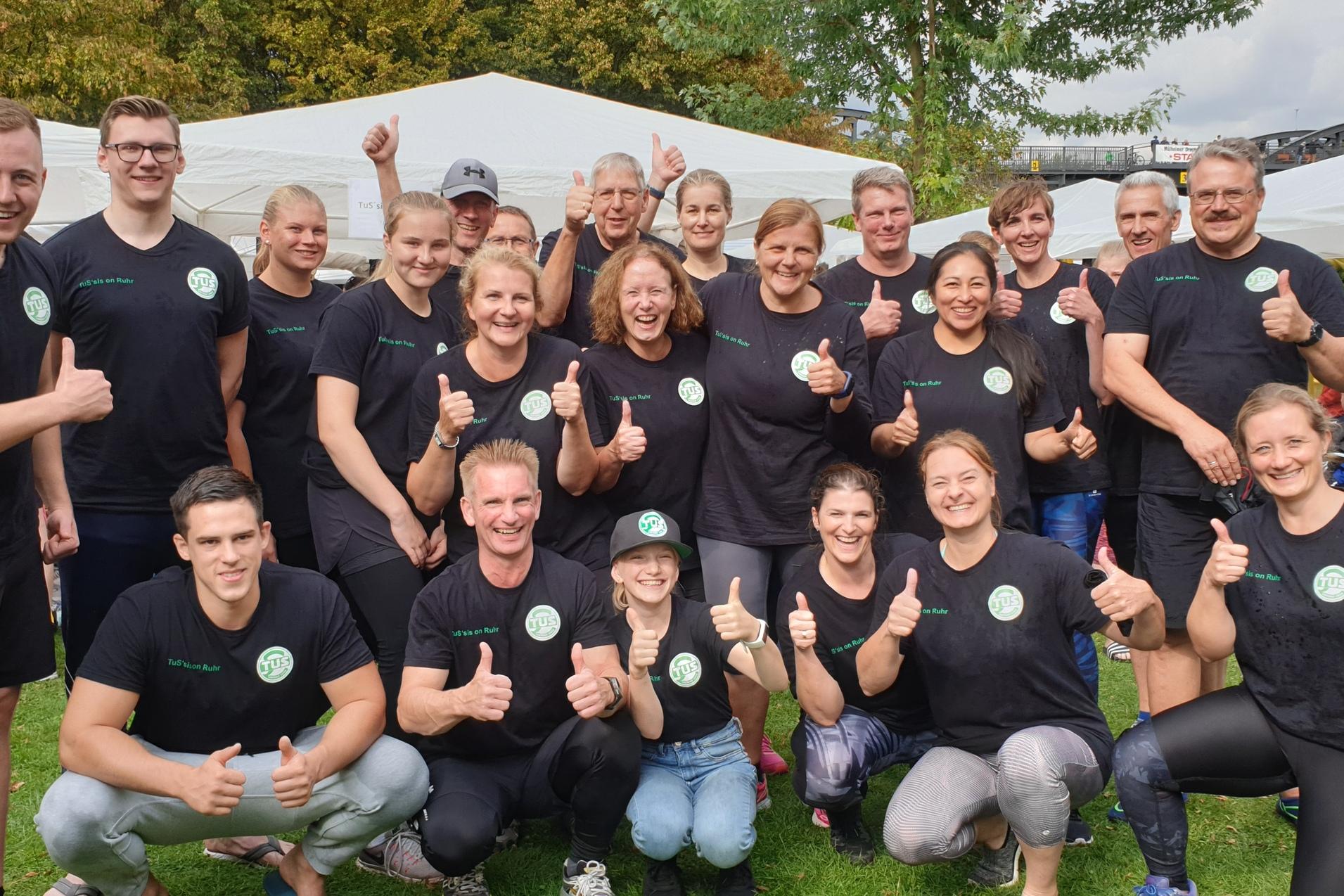 Gruppe von 16 Personen in schwarzen T-Shirts mit Daumen hoch, umgeben von Zelten und B&auml;umen im Freien.