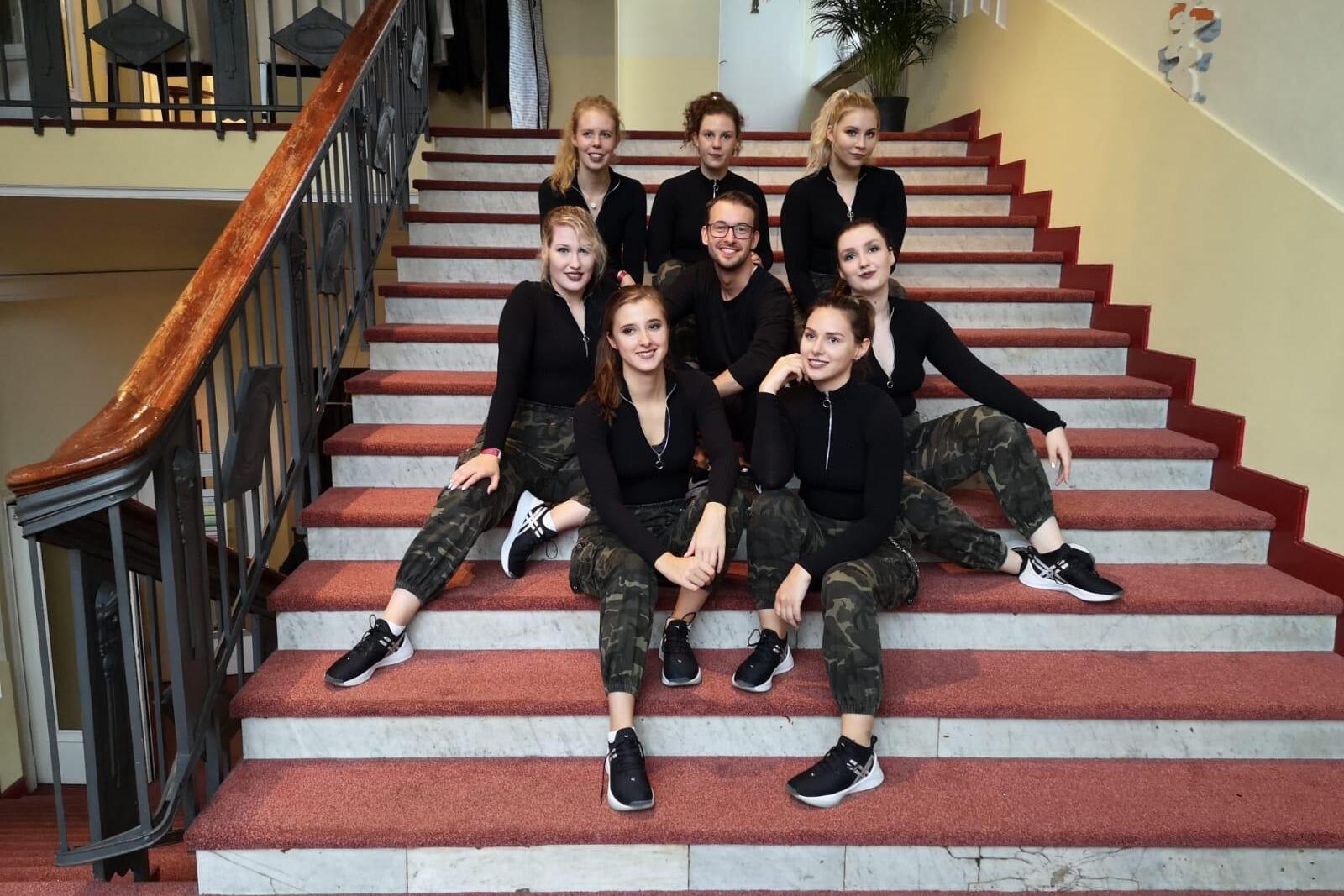 Gruppenfoto von neun Personen in schwarzer und camouflagierter Kleidung, die auf einer Treppe posieren.