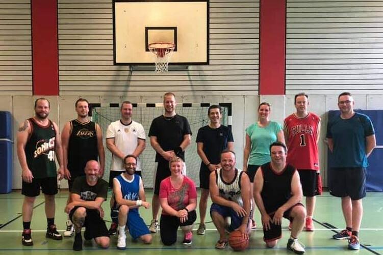 Gruppe von 14 Personen in Sportbekleidung auf einem Basketballfeld, mit einem Korb im Hintergrund.