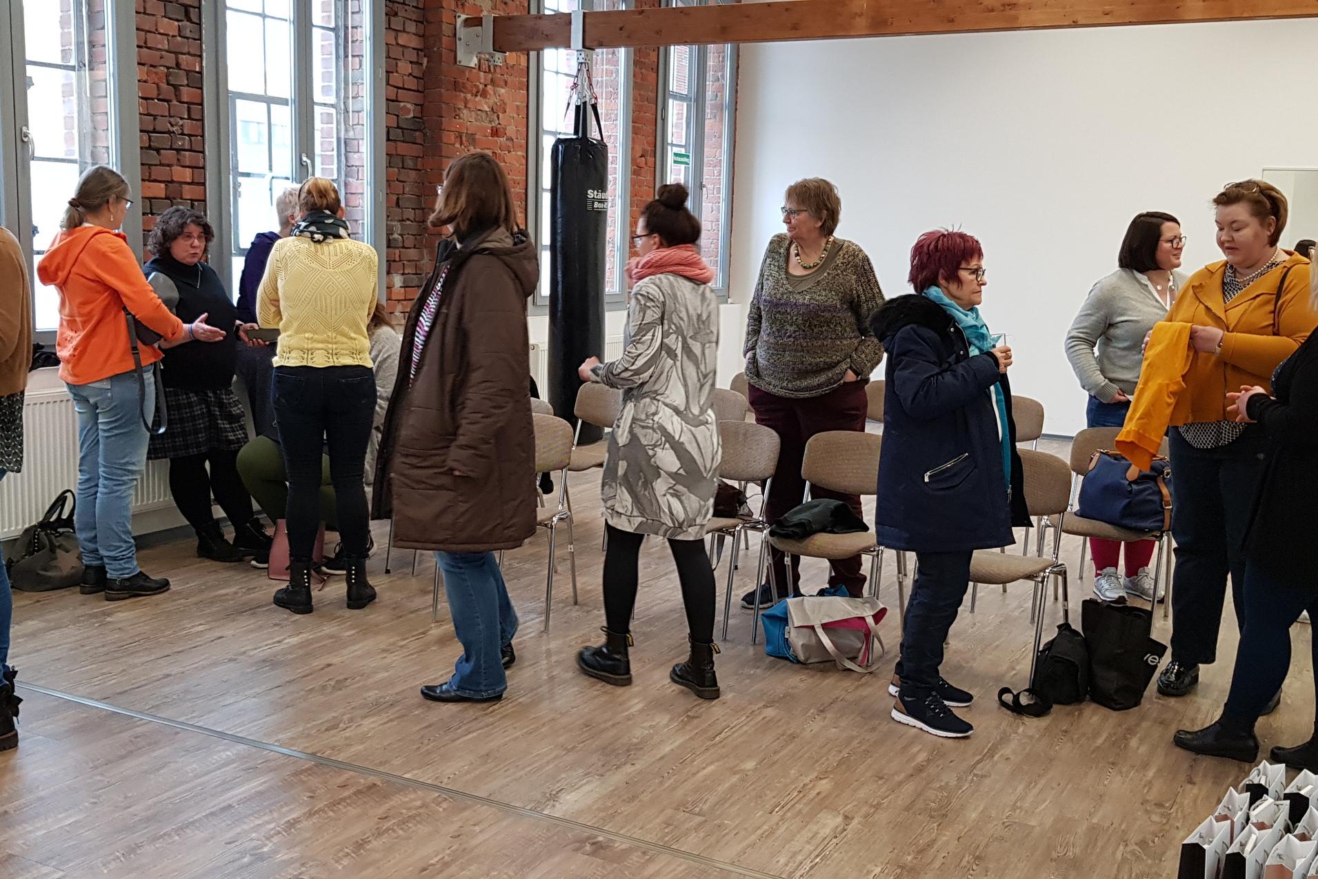 Gruppenszene in einem hellen Raum, Frauen stehen und unterhalten sich, einige sitzen auf Stühlen.
