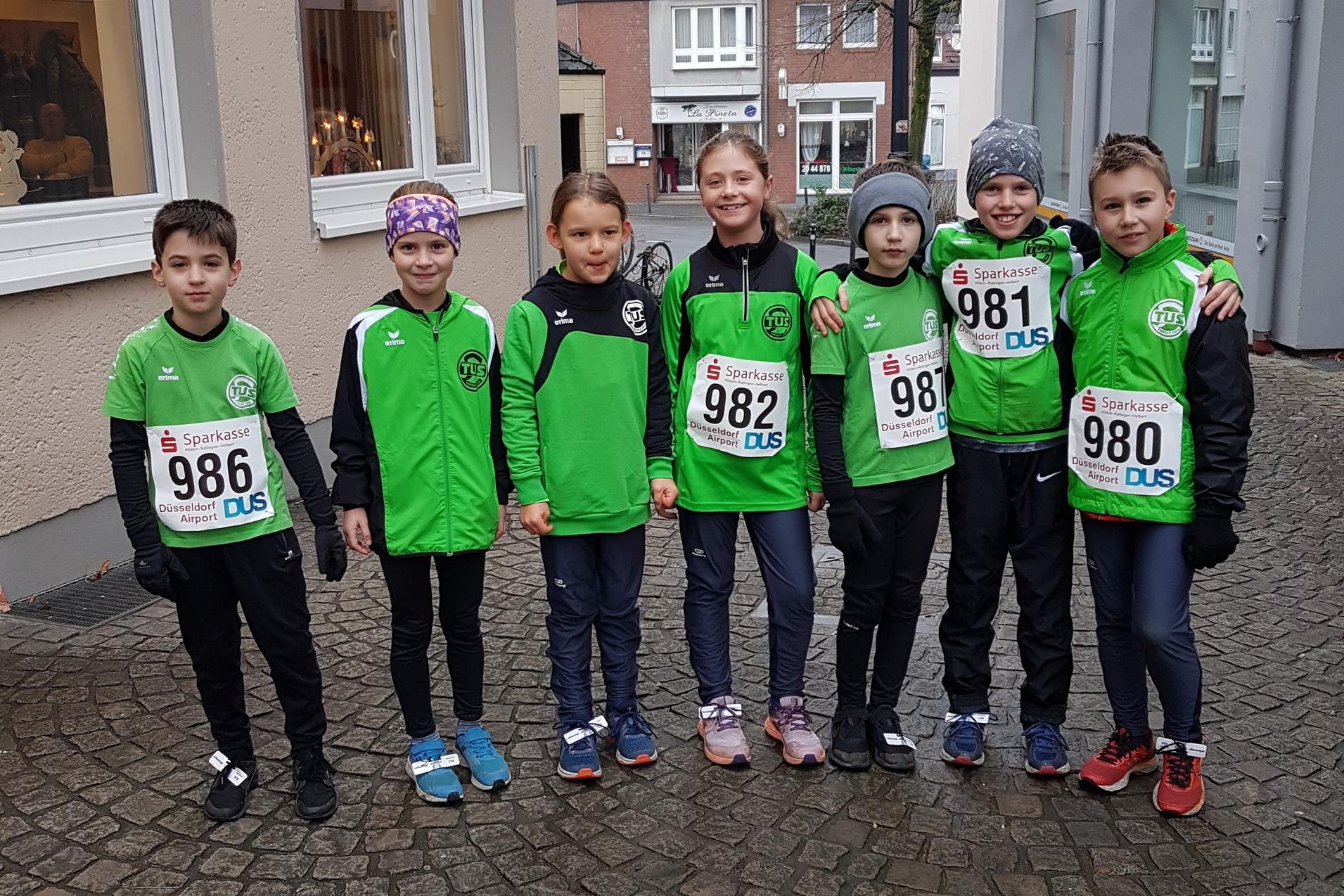 Gruppe von sieben Kindern in gr&uuml;nen Sportshirts und Startnummern auf einem Kopfsteinpflasterweg vor Geb&auml;uden.