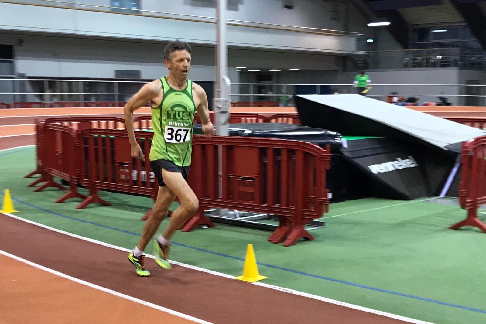 L&auml;ufer in gr&uuml;nem Trikot mit der Nummer 367 sprintet auf einer Indoor-Bahn mit roten Absperrungen und gelben Pylonen.