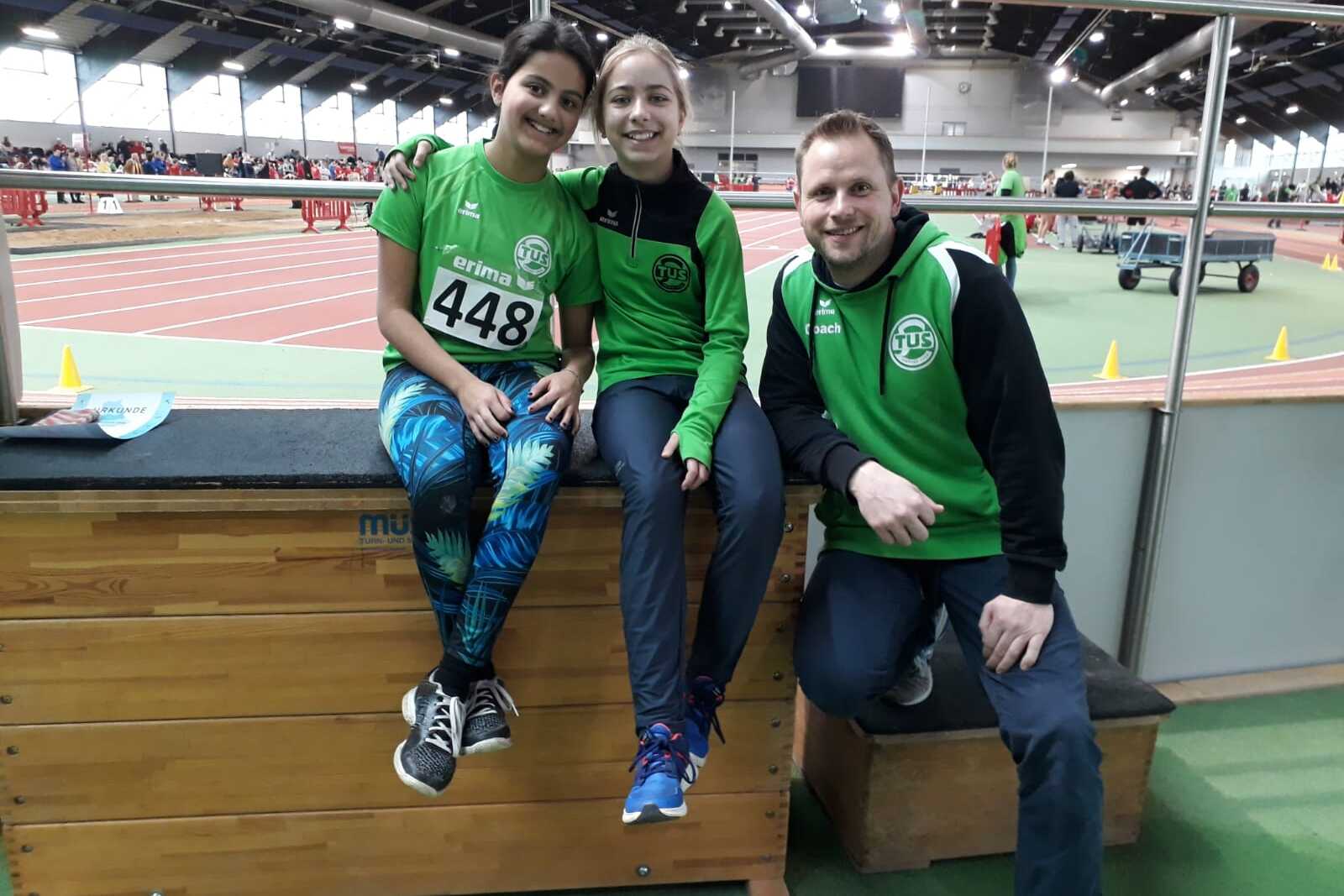 Drei Personen, zwei M&auml;dchen und ein Junge, in Sportoutfits sitzen auf einer Holzbank in einer Sporthalle.