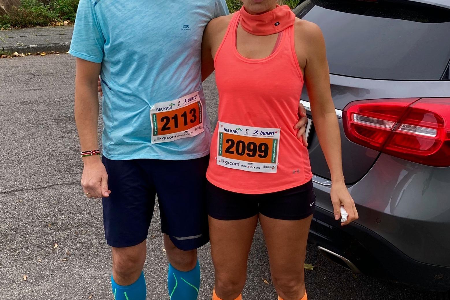 Zwei Personen in Sportkleidung stehen neben einem Auto, beide tragen Startnummern für einen Laufwettbewerb.