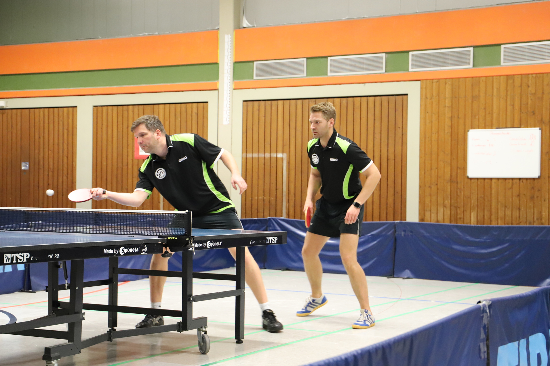 Zwei Tischtennisspieler in Uniform halten sich an einem Tischtennistisch bereit, um einen Ball zu spielen.