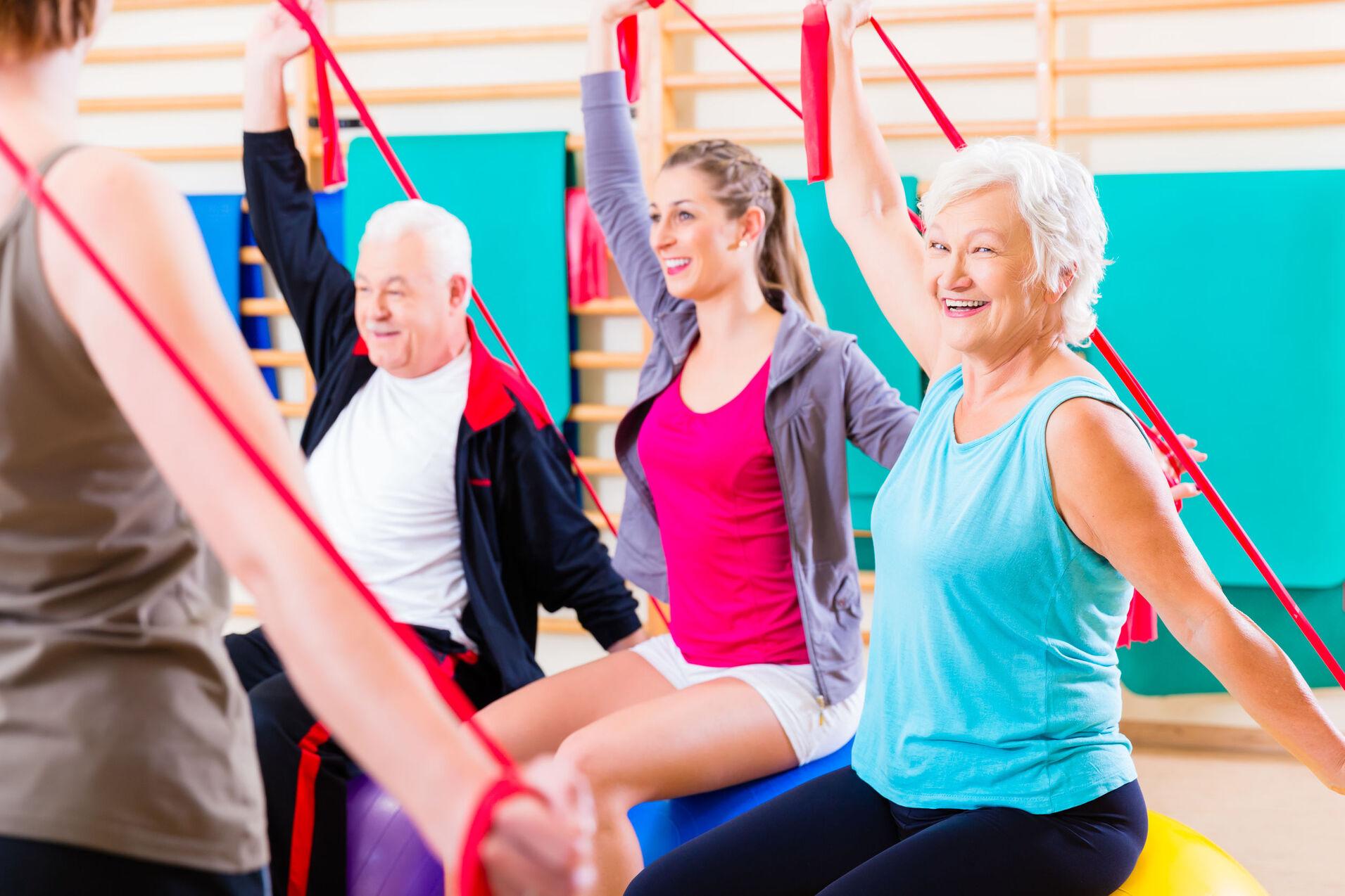 Gruppe von vier Personen, die in einem Fitnessraum mit Widerstandsbändern Übungen auf Gymnastikbällen ausführen.