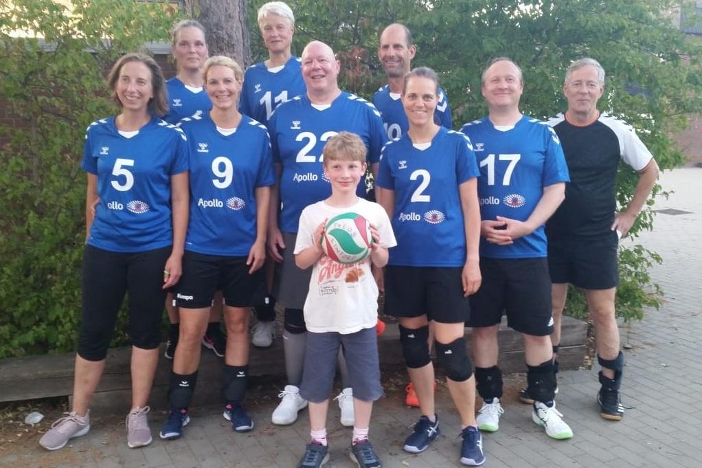 Eine Volleyballmannschaft mit zehn Spielern in blauen Trikots steht lächelnd auf einem Platz, ein Kind hält einen Volleyball.