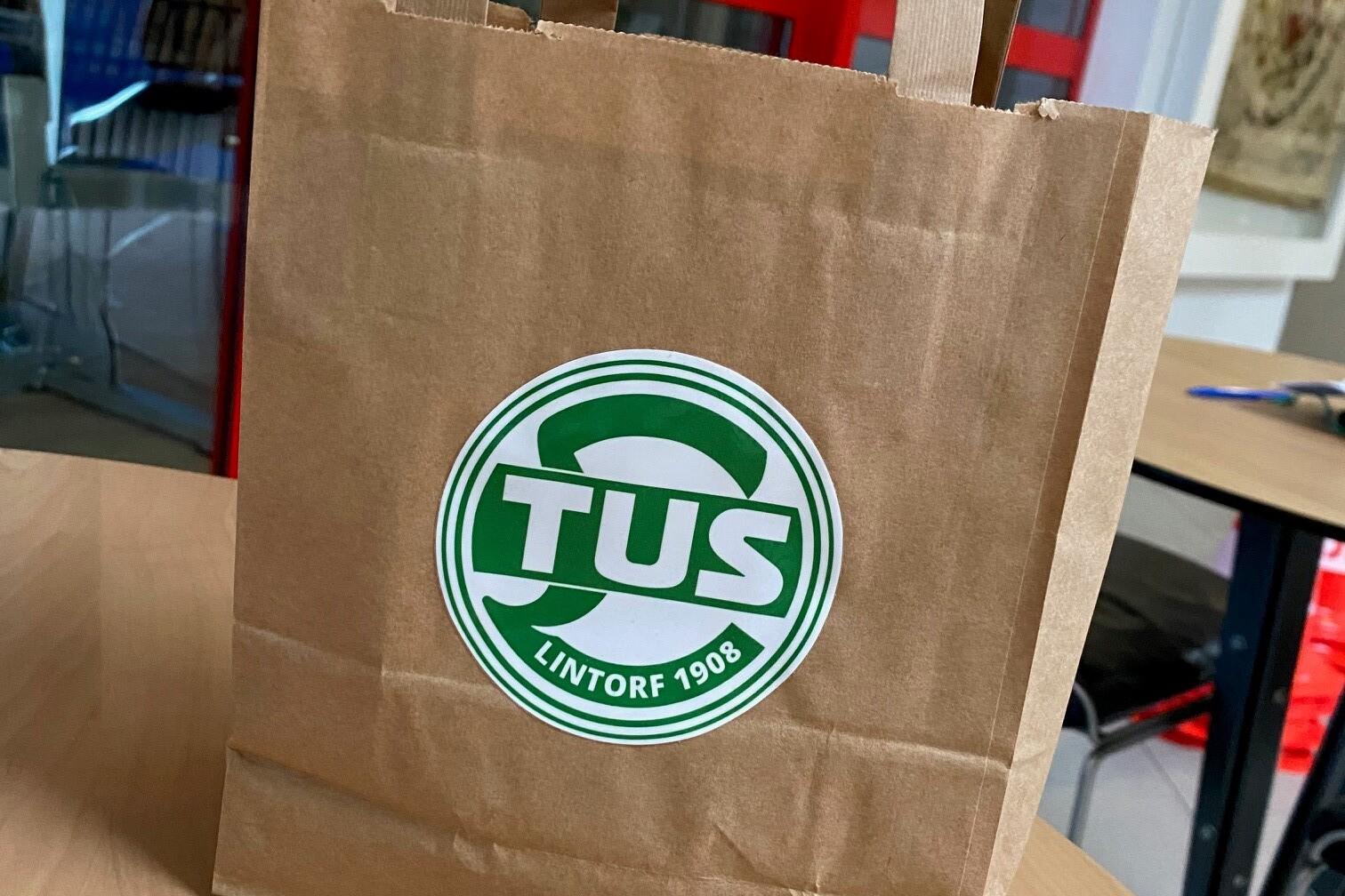 Braune Papiert&uuml;te mit dem Aufdruck &bdquo;TUS Lintorf 1908&ldquo; auf einem Tisch in einem Raum mit roten Fenstern.