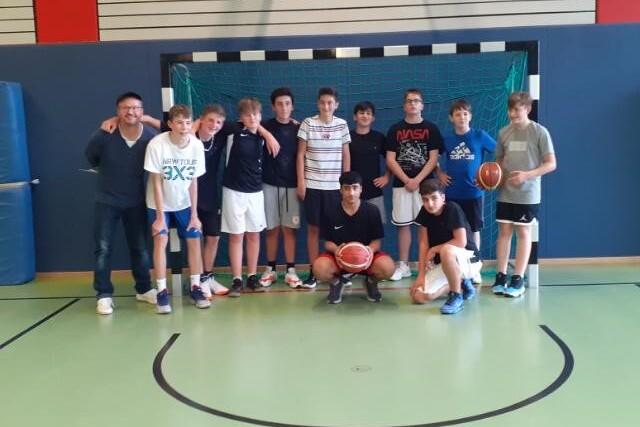 Gruppe von Jugendlichen mit Basketball im Sporthallen-Hintergrund, zwei Trainer stehen links neben den Spielern.