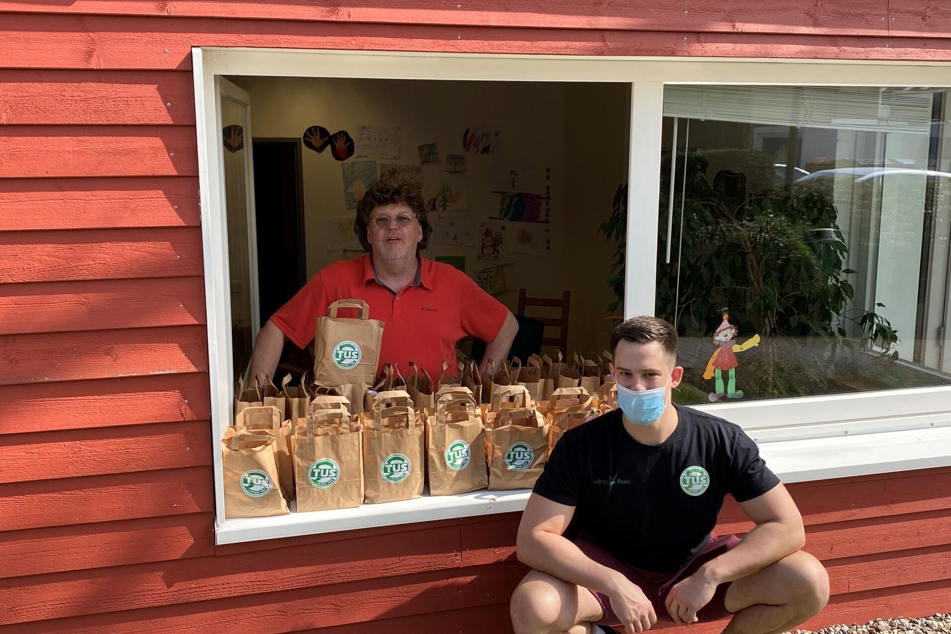 Zwei Männer stehen und sitzen vor einem Fenster, das mit Papiertüten gefüllt ist, in einem roten Holzhäuschen.