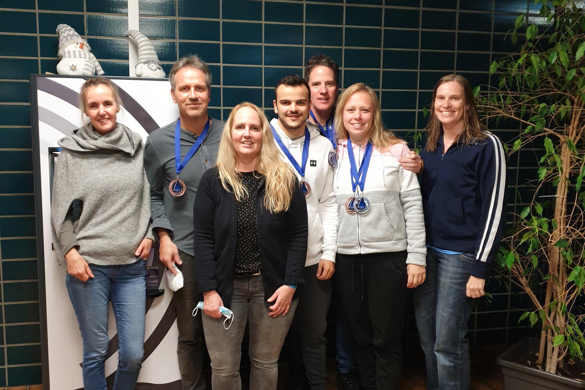 Sechs Personen mit Medaillen stehen vor einem Getränkeautomaten in einem Flur mit grünem Fliesenmuster.