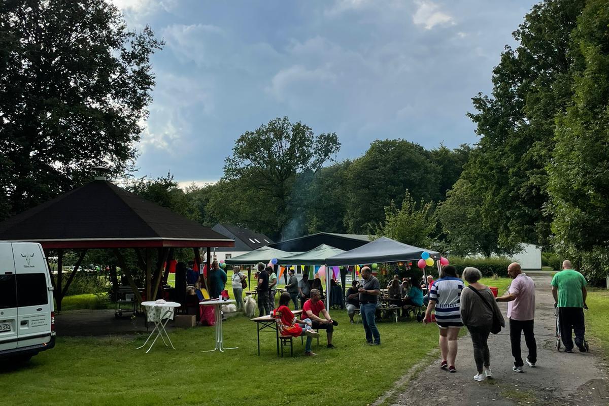 Menschengruppe in einem Park bei Festlichkeiten unter Pavillons, umgeben von Bäumen und einer wolkigen Himmel.