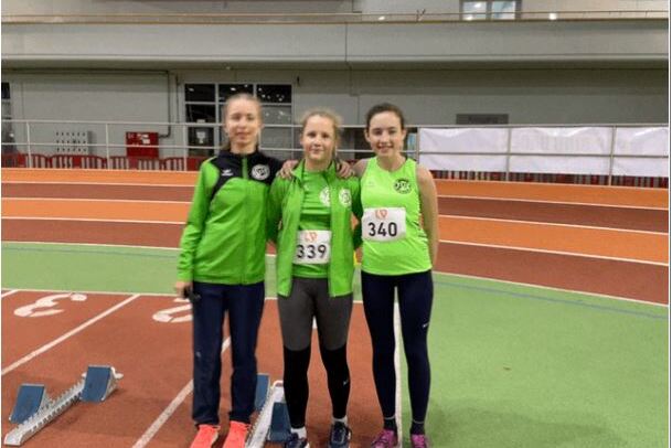 Drei Sportlerinnen in gr&uuml;nen Trikots stehen l&auml;chelnd auf einer Leichtathletikanlage mit Startbl&ouml;cken.