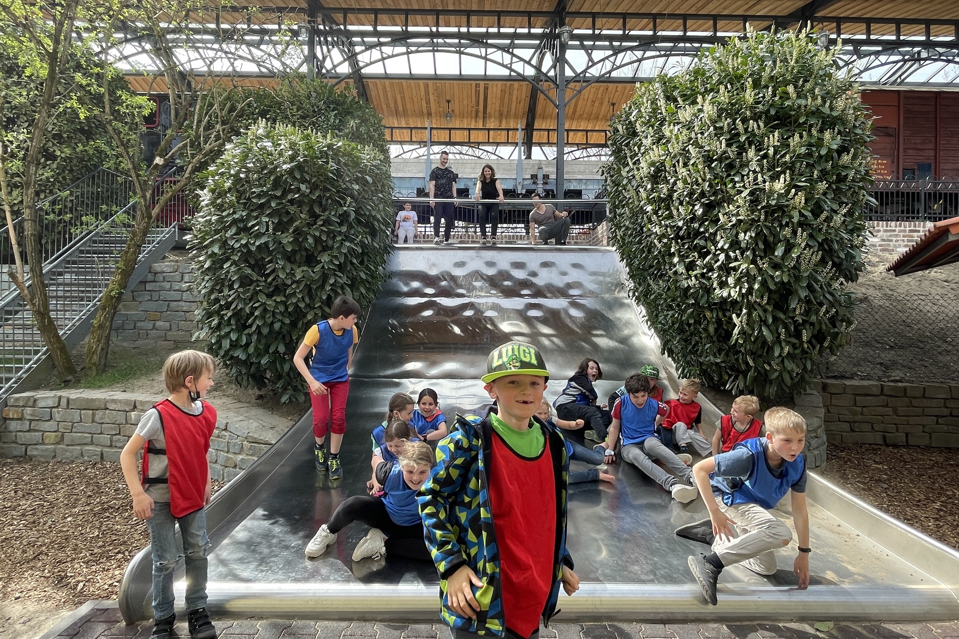 Gruppe von Kindern in roten und blauen T-Shirts spielt an einer Rutsche in einem Freizeitanlagenbereich.