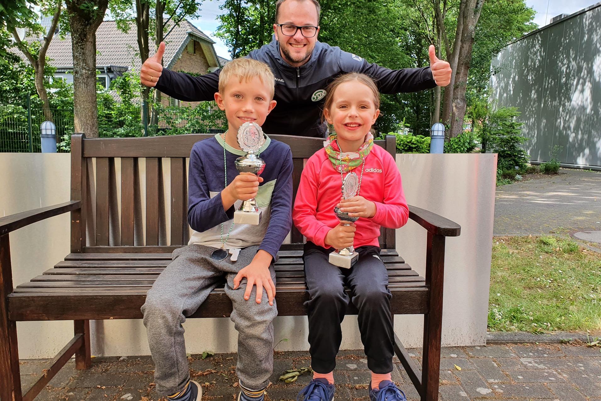 Zwei Kinder mit Pokalen sitzen auf einer Bank, während ein lächelnder Erwachsener dahinter mit Daumen hoch steht.