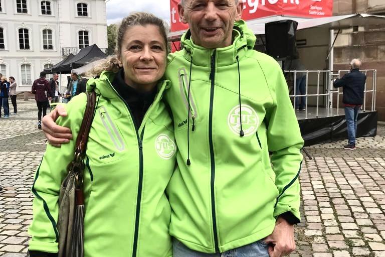 Paar in grünen Jacken umarmt sich vor einer historischen Kulisse auf einem Platz mit Kopfsteinpflaster.