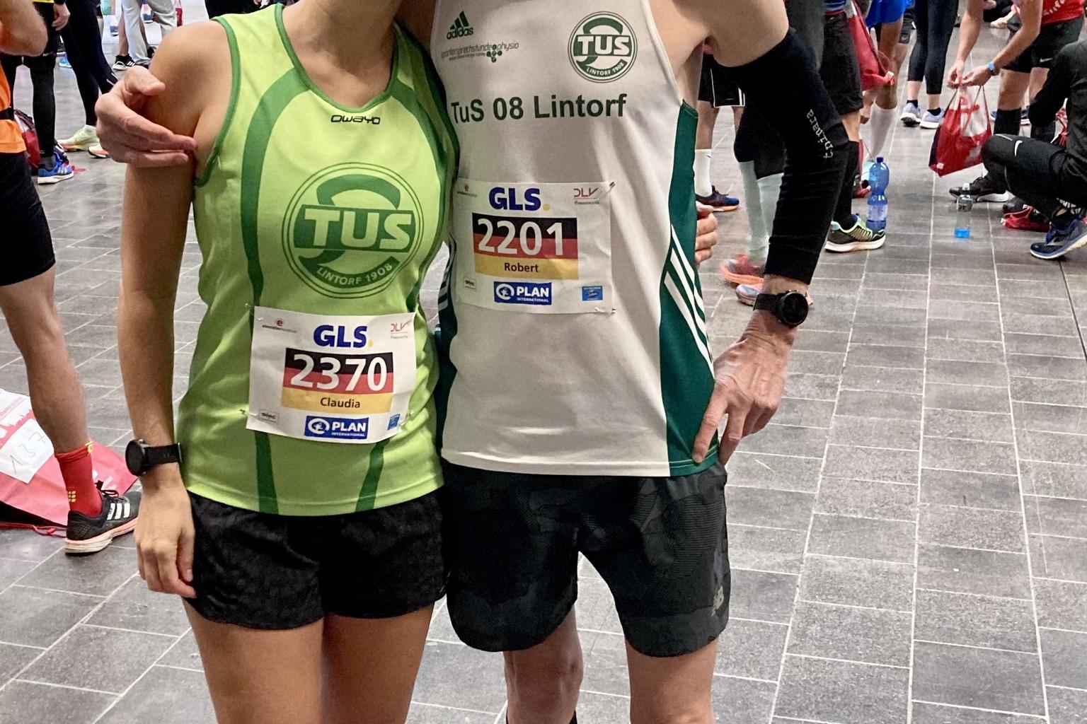 Zwei L&auml;ufer stehen in Sportbekleidung mit Startnummern in einer Halle voller Teilnehmer und Vorbereitungen.