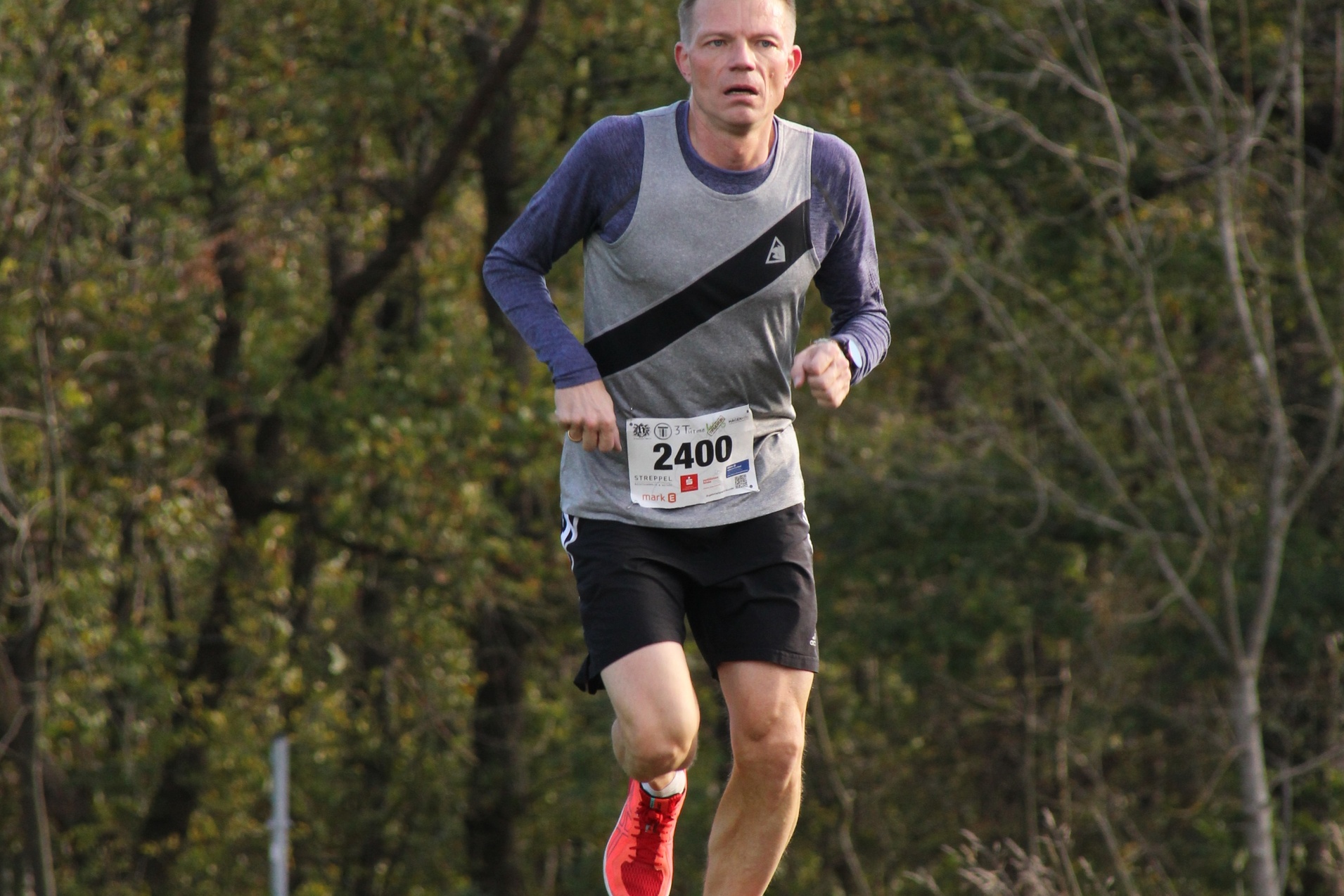 L&auml;ufer in sportlicher Kleidung mit Startnummer 2400 joggt auf einem Weg im Freien, umgeben von B&auml;umen und Gras.