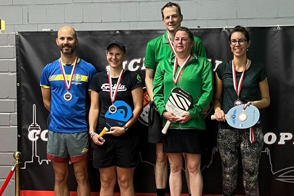 F&uuml;nf Personen stehen auf einem Siegerpodest mit Medaillen, umgeben von einer Tischtennisarena mit Werbebanner.