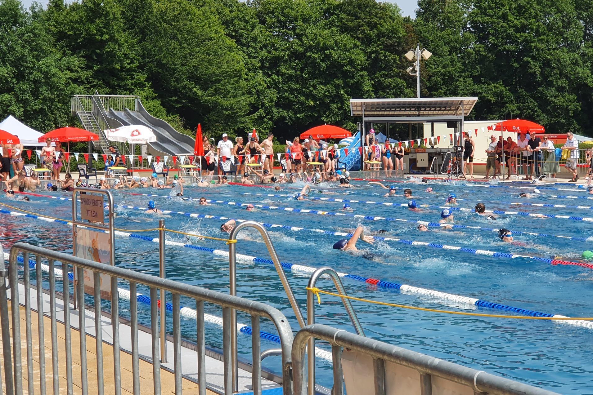 Blick auf ein belebtes Schwimmbad mit Schwimmern in einem 50-Meter-Becken und Liegestühlen unter Sonnenschirmen.
