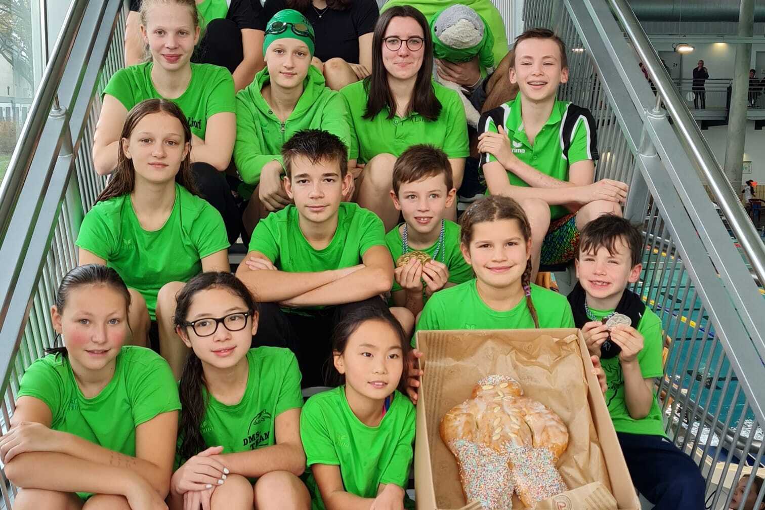 Gruppe von Schwimmern in gr&uuml;nen T-Shirts sitzt auf Treppe, eine Kiste mit Geb&auml;ck befindet sich im Vordergrund.