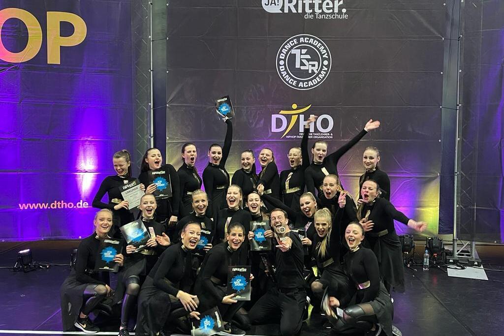Gruppe von T&auml;nzerinnen in schwarzen Outfits h&auml;lt Pokale und jubelt auf einer B&uuml;hne mit buntem Hintergrund.