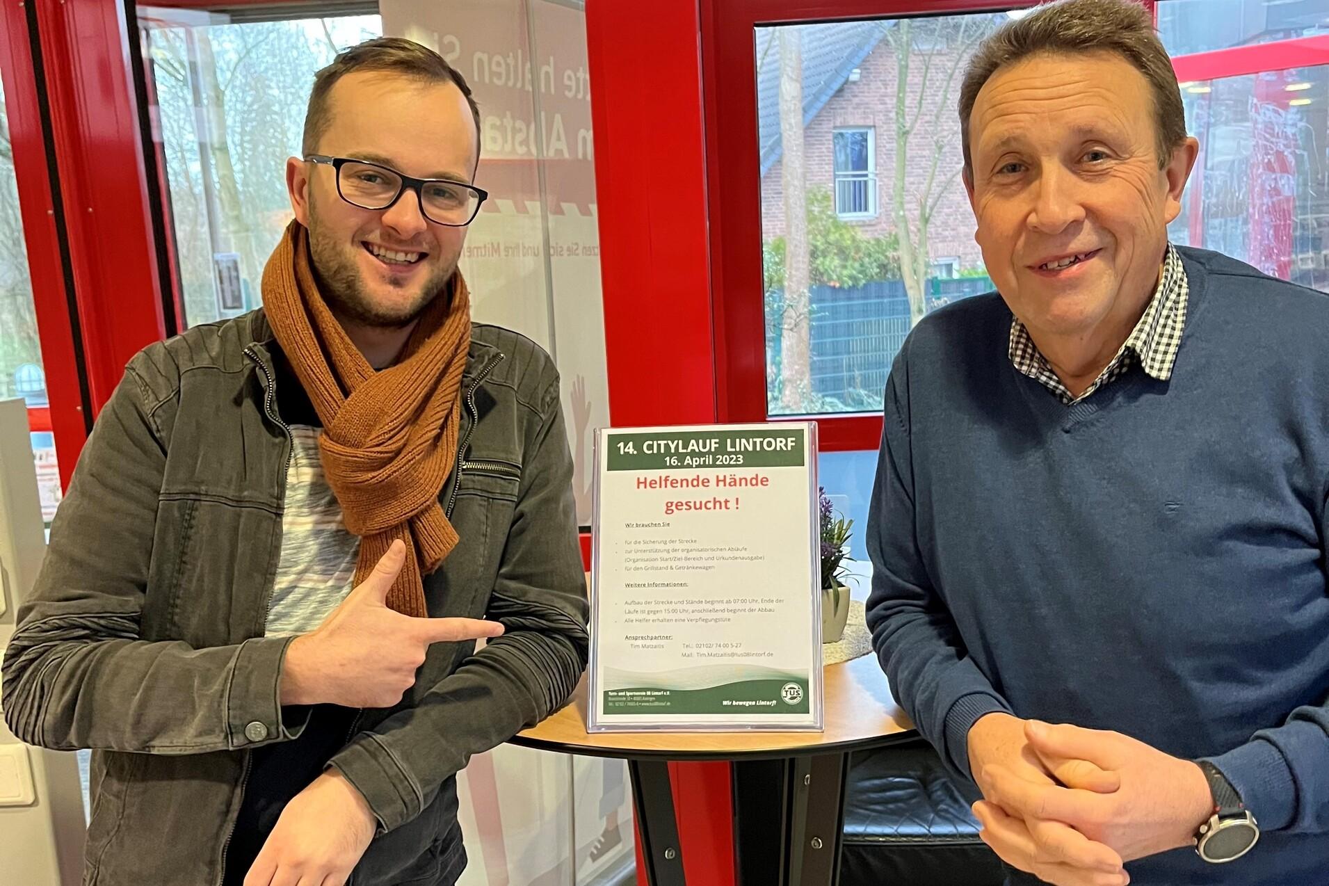 Zwei Männer stehen lachend vor einem Informationsschild mit dem Text „Helfende Hände gesucht“ für den Stadtlauf.