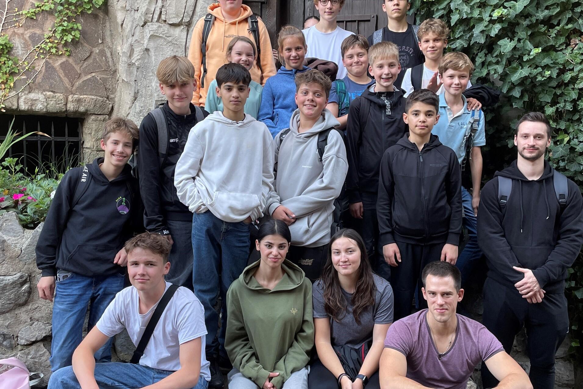 Gruppe von 20 Jugendlichen sitzt und steht vor einer begr&uuml;nten Wand und einer Holzt&uuml;r, l&auml;chelnd in die Kamera.