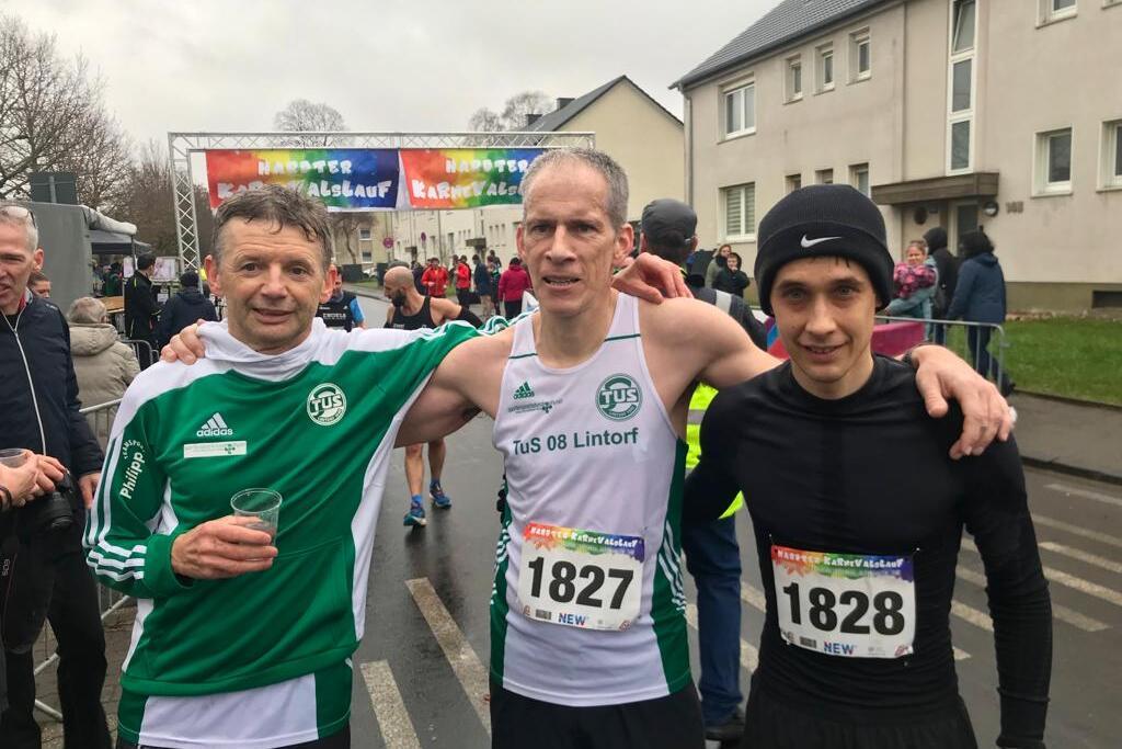 Drei M&auml;nner in Laufbekleidung stehen l&auml;chelnd vor einer Laufveranstaltung, im Hintergrund Zuschauer und ein Zielbanner.