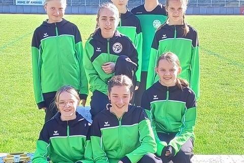 Gruppe von acht Jugendlichen in gr&uuml;nen Sportjacken, l&auml;chelnd auf einem Rasenplatz, umgeben von B&auml;umen.