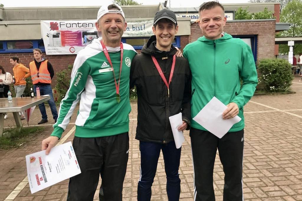 Drei Männer in Sportkleidung stehen lächelnd zusammen, halten Urkunden und Medaillen nach einem Wettkampf.