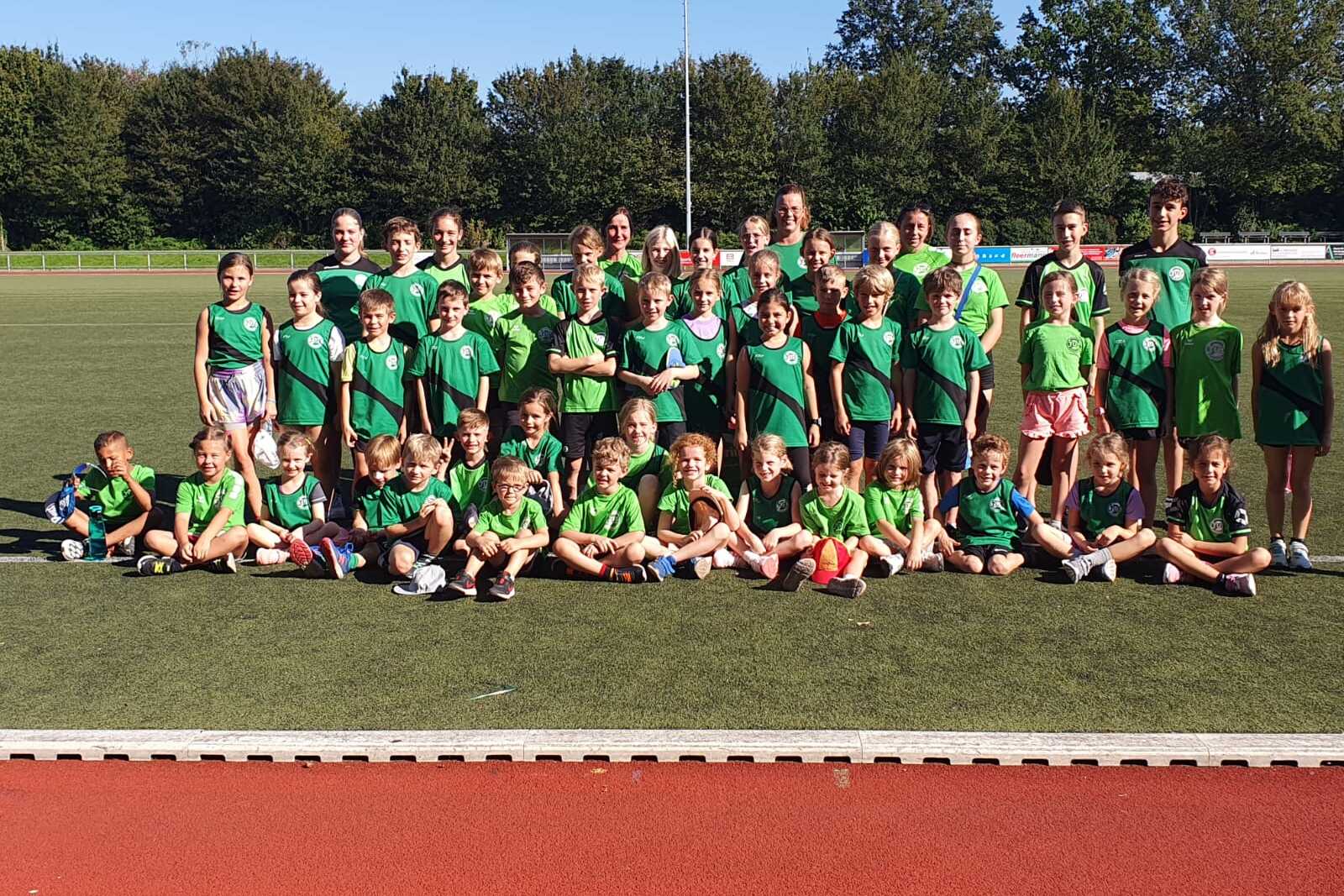 Gruppe von etwa 50 Kindern und Jugendlichen in gr&uuml;nen T-Shirts, posierend auf einem Sportplatz mit B&auml;umen im Hintergrund.