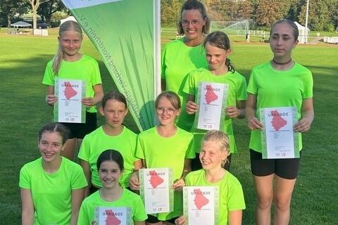 Gruppe von acht Mädchen in neonfarbenen T-Shirts, die stolz Urkunden auf einem Sportplatz halten.