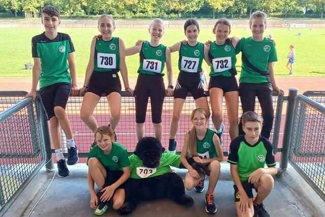 Gruppe von zehn Kindern in Sportkleidung mit Startnummern, posierend mit einem Stofftier auf einem Sportgelände.