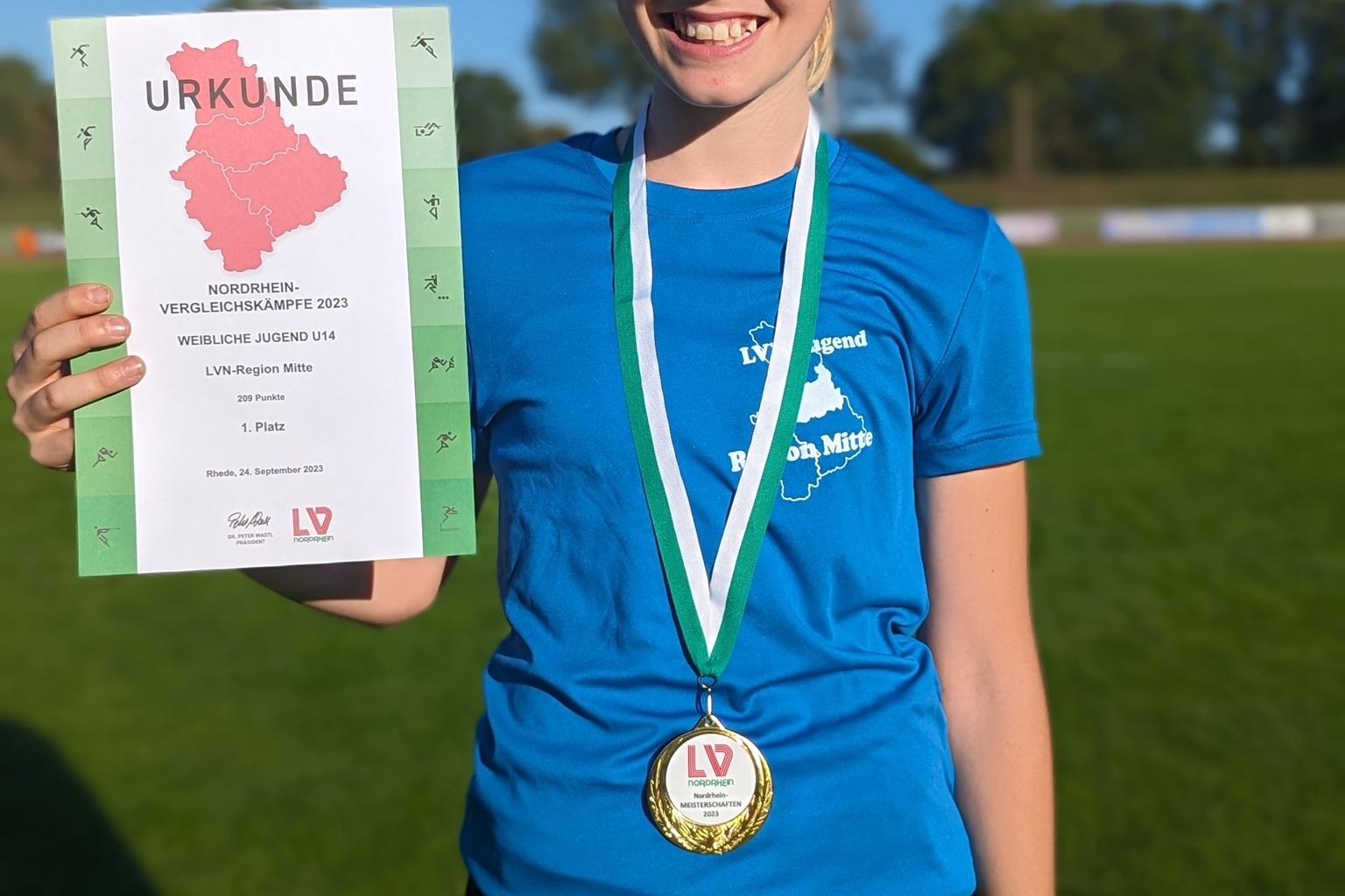 Mädchen mit Medaille und Urkunde auf einem Sportplatz, lächelnd in blauer Sportkleidung, hinter ihr Bäume und ein Fußballfeld.
