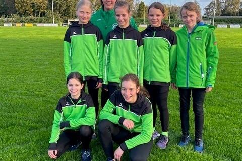 Gruppe von sieben jungen Sportlerinnen in gr&uuml;nen Trainingsjacken auf einer Wiese, l&auml;chelnd posierend.